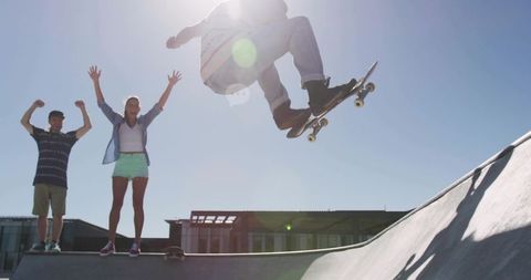 Skateboarder soaring over ramp while friends cheer at urban skatepark with lens flare