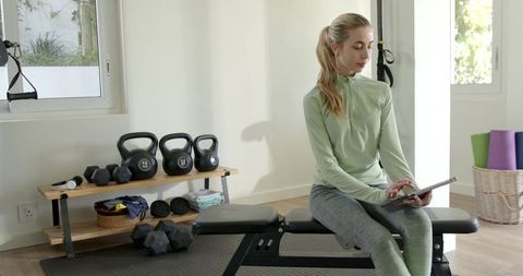 Young woman sitting on weight bench using tablet in bright minimalist home gym