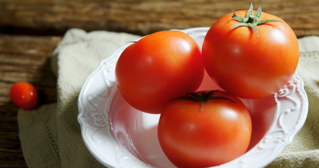 Three fresh red tomatoes sit in a white ornate bowl atop a rustic wooden table. A single small tomato is placed beside the bowl. Perfect for use in culinary blogs, healthy lifestyle articles, or vegetable gardening content.