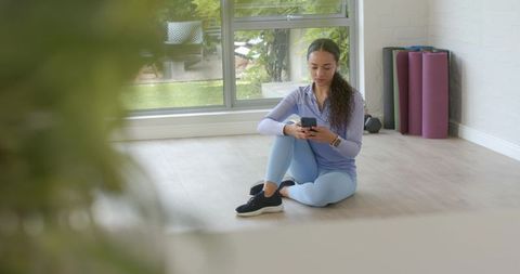 Checking phone after home workout: woman in lavender activewear sitting on sunlit floor