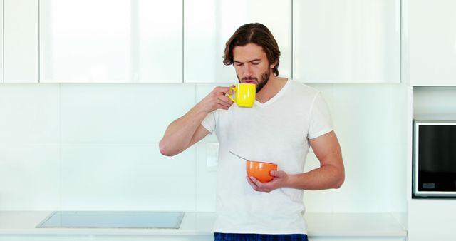 Young man drinking coffee and eating breakfast in a modern white kitchen. Ideal for advertising kitchenware, breakfast foods, or lifestyle products. Can be used for promoting morning routines, casual home living, or healthy eating habits.