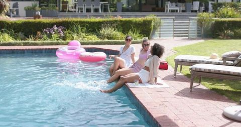 Diverse female friends sitting poolside dipping feet splashing and laughing on summer day