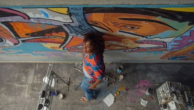 High-angle muralist standing painting vibrant street art wall with ladder and paint cans