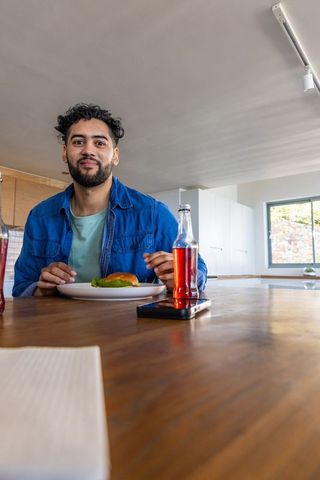 Young man sitting at modern kitchen island eating sandwich with soda and smartphone nearby