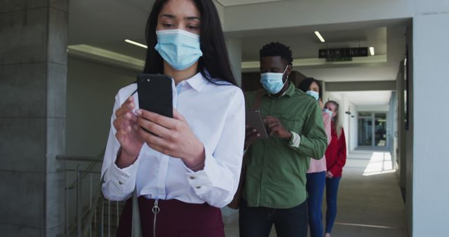 Several individuals stand in a line, spacing apart from each other while using their smartphones. They wear face masks, emphasizing modern safety practices and technology use during a pandemic. Useful for themes on health guidelines, technology in daily life, or social behavior during COVID-19.