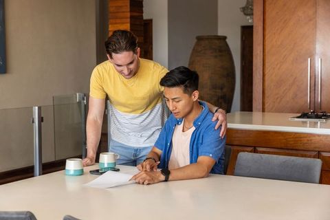Diverse male friends signing contract at home kitchen table, reviewing paperwork together