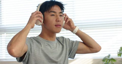Asian man adjusting white headphones listening at home with smartwatch and blinds