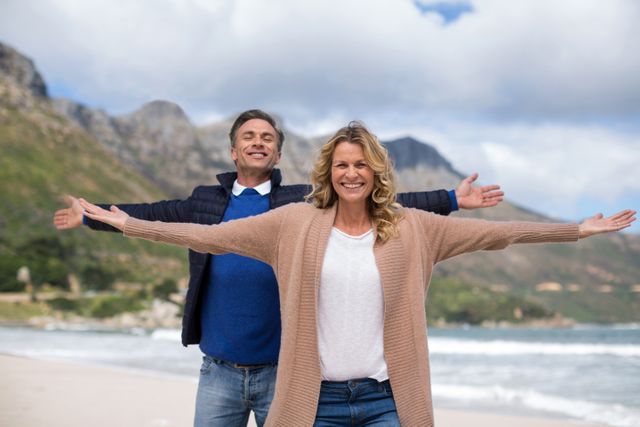 Mature couple standing on a beach with arms outstretched, smiling and enjoying the moment. Mountains and ocean in the background. Ideal for travel brochures, retirement planning advertisements, wellness and lifestyle blogs, and romantic getaway promotions.
