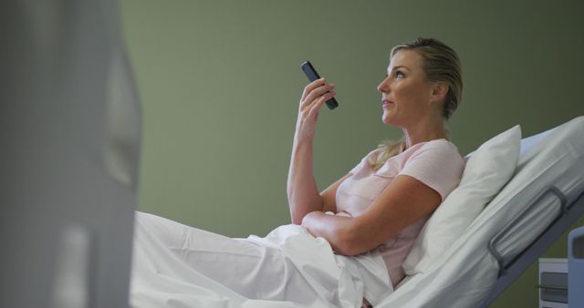 Woman lying in hospital bed, holding a remote control in a hospital room. Suitable for use in healthcare and medical advertisements, patient care articles, and hospital focused publications.