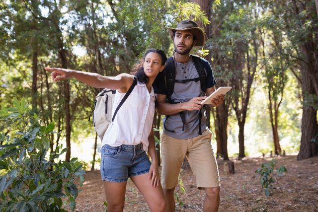 Couple exploring forest with tablet and backpack. Woman pointing while man holds tablet, indicating navigation or direction. Ideal for use in travel blogs, adventure promotions, hiking gear advertisements, and nature exploration articles.