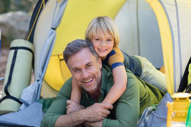 Smiling little boy lying on father in tent at campsite