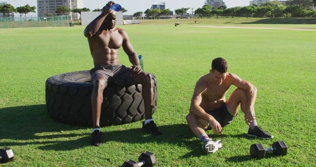 Two athletes sitting on a grassy field after a strenuous workout session. One athlete is sitting on a large tractor tire, pouring water on his head, while the other is sitting on the ground next to dumbbells. Ideal for fitness blogs, exercise equipment advertisements, and articles on outdoor training routines.