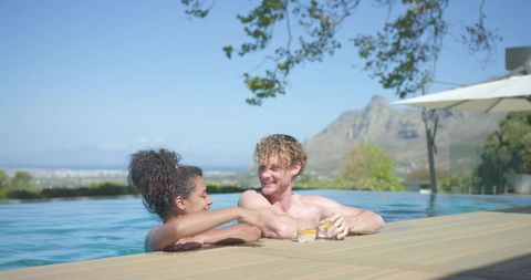 Couple relaxing in infinity pool on scenic coastal terrace with cool drinks, sunshine