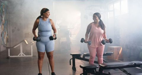 Diverse women lifting dumbbells and smiling during strength workout in bright gym