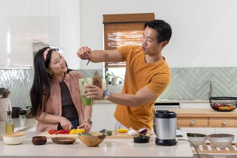 Asian couple preparing green smoothie while chopping vegetables on modern kitchen island