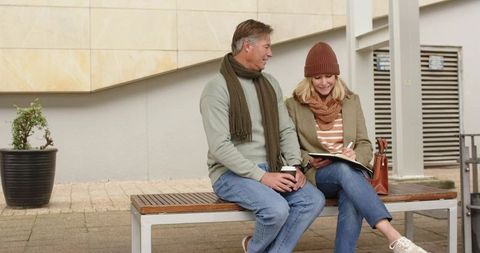 Middle-aged couple sharing coffee while woman writing in notebook on city bench