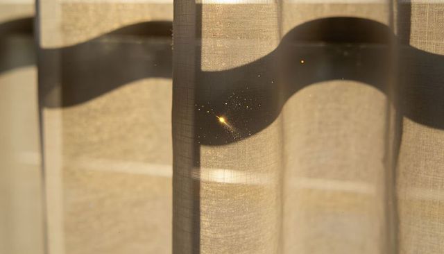 Sunlit sheer curtain with vertical pleats and golden dust sparkles, warm fabric texture and soft sha