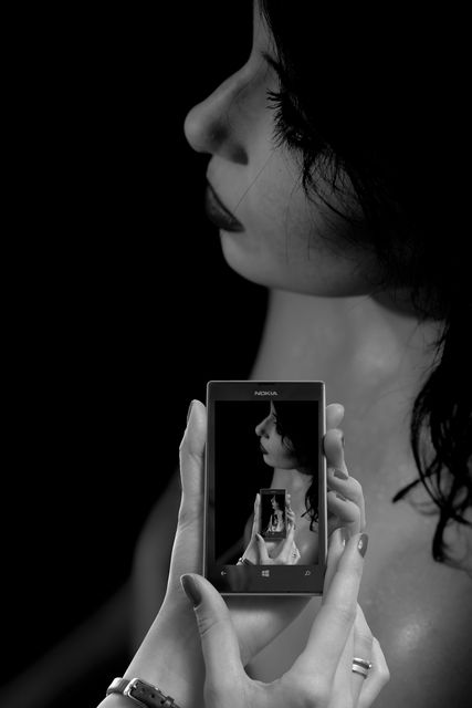 A contemplative woman holding a smartphone captures her reflection in a mirror. Black and white format adds a dramatic effect. Suitable for themes of self-reflection, technology in daily life, and introspection. Ideal for use in blogs, social media posts, and mental health awareness campaigns.