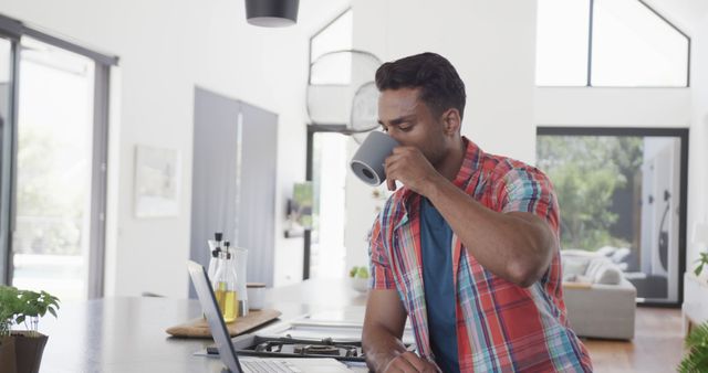 Young man in plaid shirt drinking coffee working on laptop in modern kitchen - Download Free Stock Images Pikwizard.com