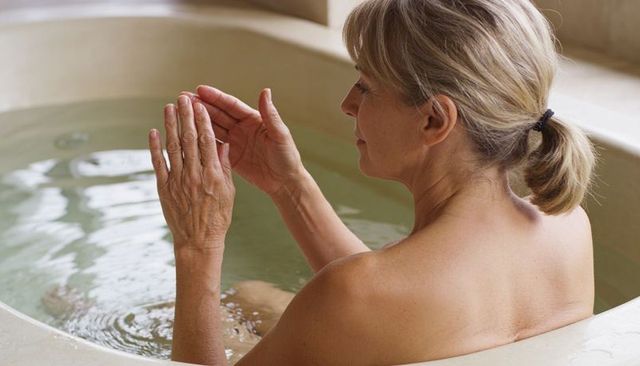Mature woman soaking in circular stone tub cupping water at private spa, peaceful self-care