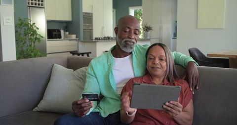 Mature diverse couple sitting on sofa using tablet and credit card for online shopping