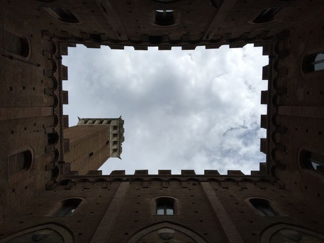 This photo captures an upward view of a historic brick tower framed by a courtyard, with a cloudy sky in the background. Ideal for use in travel brochures, historical architecture presentations, or cultural articles highlighting medieval structures and tourism in Italian cities. The contrast between the architectural detail and the sky adds visual interest.