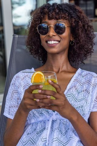 Smiling african american woman wearing sunglasses sipping refreshing citrus drink on sunny patio
