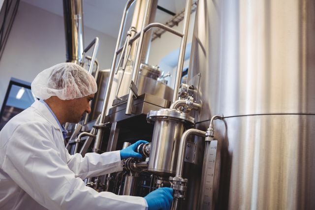 Manufacturer in protective gear working at stainless steel storage tanks in a brewery. Ideal for use in articles about industrial processes, brewing industry, manufacturing, and professional work environments.