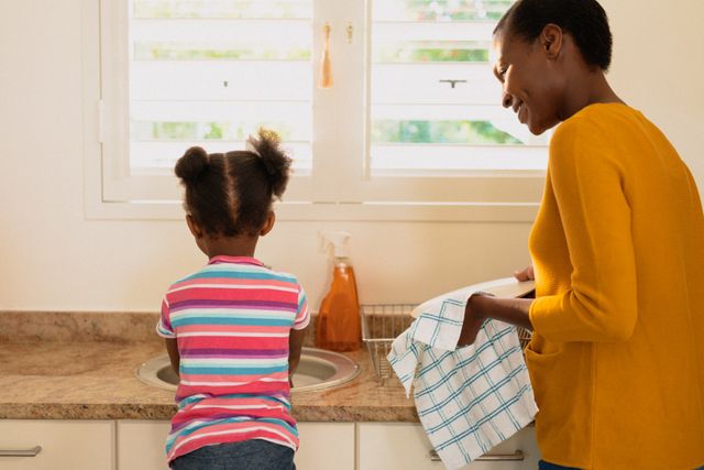 African American mother and daughter washing dishes together in a bright kitchen. The mother is smiling while drying dishes, and the daughter is standing on a stool at the sink. This image can be used to depict family bonding, domestic chores, and parenting. It is ideal for articles or advertisements related to family life, household products, and parenting tips.