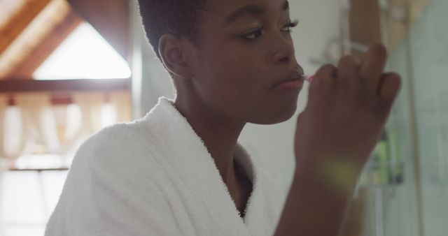 Young woman brushing her teeth in a modern bathroom, integral part of morning hygiene routine. Wearing a white bathrobe, she stands next to a mirror and sink, focusing on tooth cleaning. Useful for topics related to personal care, health, daily habits, morning routines, and cleanliness promotions.