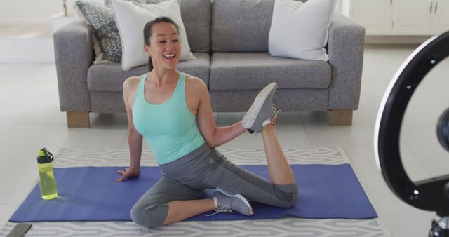This photo shows a smiling woman practicing yoga in her living room while following an online class setup on a ring light. It can be used for websites or articles related to home fitness routines, online exercise classes, wellness blogs, or social media posts promoting a healthy lifestyle.