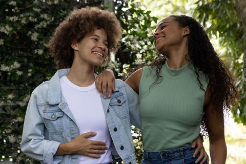 Two african women laughing and embracing in sunlit garden with denim, rings, curly hair fashion