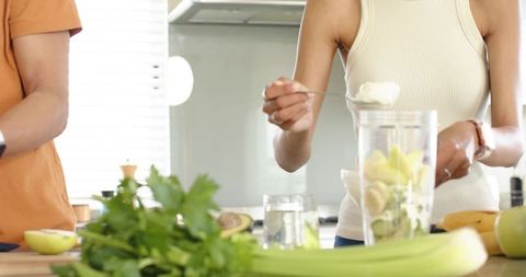 Diverse couple making green smoothie with avocado, banana, celery and parsley in kitchen