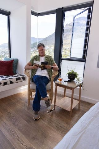 Mature man reading in sunlit bedroom nook with wicker chair and mountain view