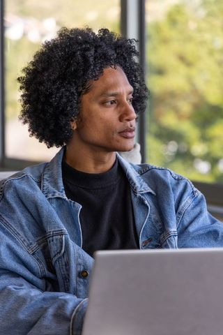 Contemplating african american man using laptop in sunlit home workspace wearing denim jacket