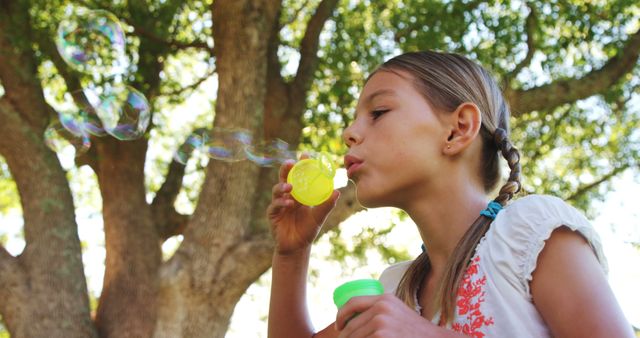 A young girl is blowing bubbles in a park, with copy space. She appears focused and delighted by the floating soap bubbles on a sunny day.