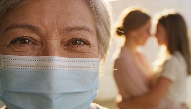 Close-up senior woman wearing surgical mask with family members embracing in warm backlight