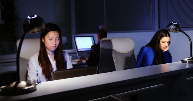 Overworked employees at their desks with computer screens, working late into the night in a dimly lit office. Ideal for articles on work-life balance, stress, and office culture.