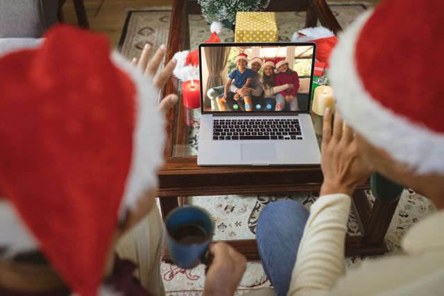 Diverse couple wearing santa hats video calling a happy family for holiday celebration. Tablet displaying image of joyful family members smiling and wearing santa hats. Cozy living environment with holiday decor and candles. Ideal for holiday communication, remote family connections, and festive season marketing.