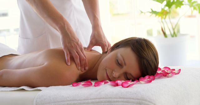 A young Caucasian woman enjoys a relaxing massage in a serene spa environment, with copy space. Her content expression and the presence of rose petals add to the tranquil ambiance of a wellness retreat.