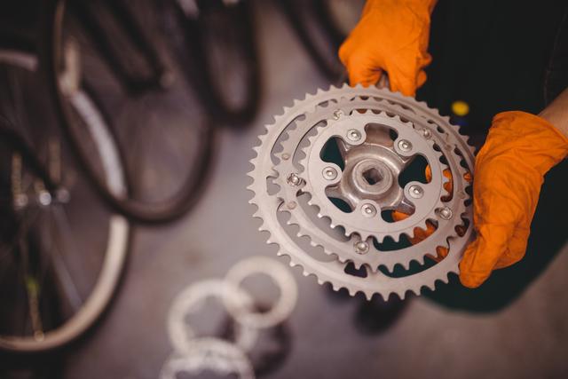 Mechanic holding bicycle gear in workshop, wearing orange gloves. Ideal for illustrating bicycle repair, cycling maintenance, mechanic workshops, and bike parts businesses.