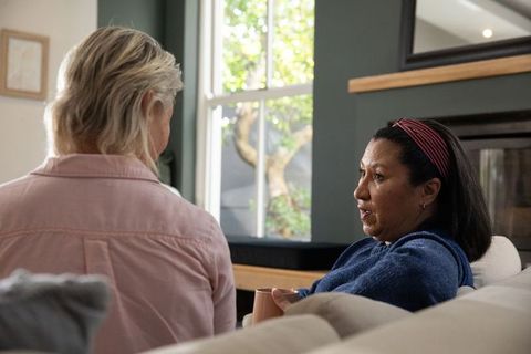 Senior friends having heartfelt conversation on sofa in sunlit living room with coffee mug