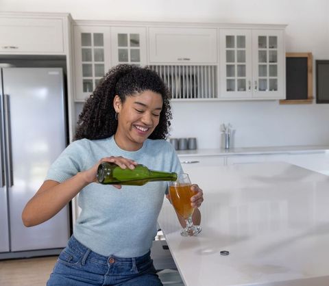 Smiling woman pouring amber ale from green bottle at bright minimalist kitchen island