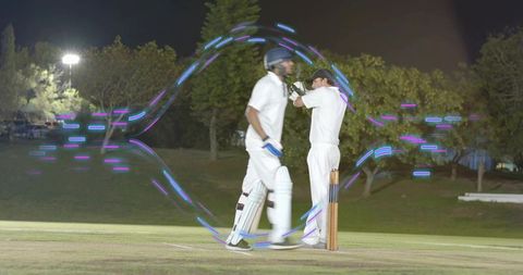 Cricket players walking on night pitch with neon light trails circling stumps