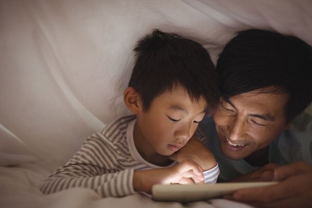 Father and son bonding while using a digital tablet under a blanket in a cozy bedroom setting. Ideal for use in articles or advertisements about family time, technology in parenting, bedtime routines, or childhood learning. Perfect for illustrating concepts of togetherness, parent-child relationships, and modern family life.