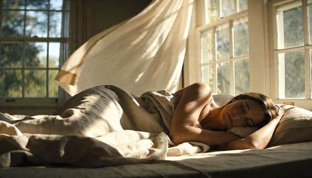 Serene woman sleeping in sunlit bedroom with flowing curtains and natural linen bedding