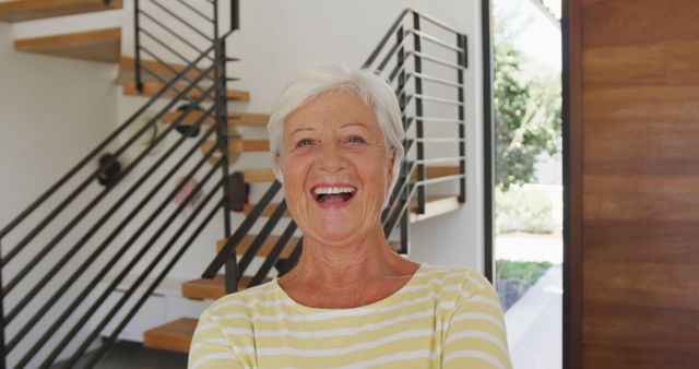 This image showcases a joyful elderly woman standing indoors in front of a modern staircase. Great for illustrating positivity, aging, retirement, happiness, and indoor lifestyles. Suitable for use in health and wellness campaigns, retirement planning, senior living advertisements, and greeting cards.