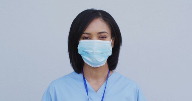 Healthcare professional dressed in light blue scrubs and wearing a surgical mask standing against a plain background. Can be used in contexts related to medical staff, safety protocols during the pandemic, healthcare services, frontline workers, and medical recruitment.