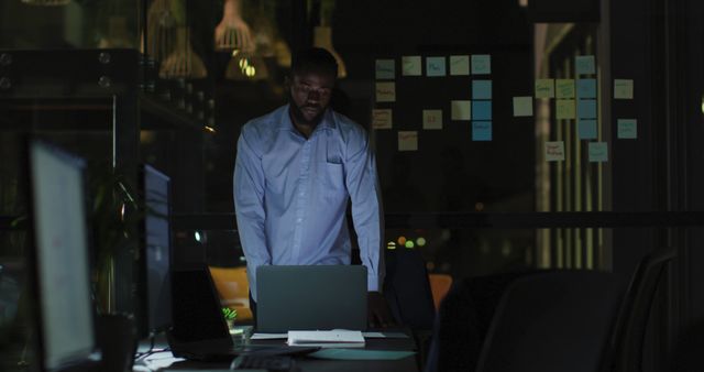 Image of tired african american businessman working late, leaving laptop and office desk at night. Business, communication, inclusivity and flexible working concept digitally generated image.