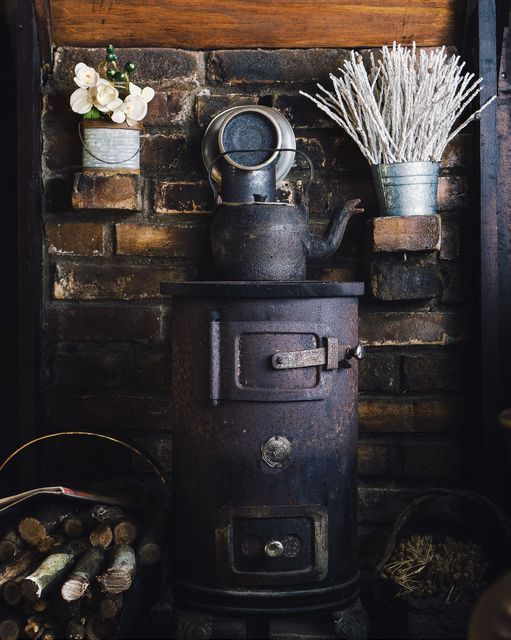 This scene captures a rustic wood-burning stove with a vintage tea kettle on top, surrounded by flowers in small pots. The stove against a brick wall adds a cozy and warm atmosphere, ideal for themes of rustic home decor, countryside living, and vintage aesthetics. Perfect for use in lifestyle blogs, home improvement magazines, or advertisements for rustic-themed products.